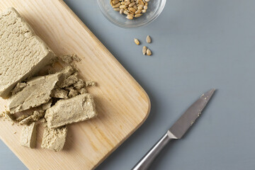 Crumbled sunflower halva lies with a metal knife on a cutting wooden board on a gray background. Concept of vegetarian sweets. Horizontal orientation. Copy space. Top view.
