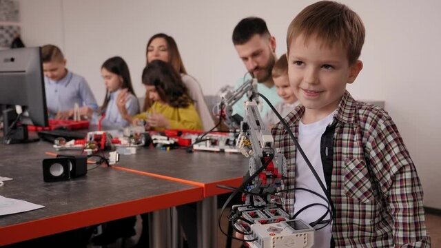 Portrait of a happy schoolboy with a robot in robotics lessons at school, in the background children with their parents are sitting at computers and assembling a constructor.