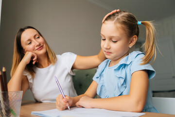 Close-up of cute primary daughter doing homework with young mother sitting at home table by window. Skilled smiling female tutor teaching elementary school girl helping with lesson.