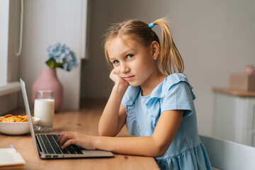 Side view of exhausted elementary child girl using laptop sitting at home table with snack by window, looking at camera. Portrait of little primary kid chatting having distance studying online.