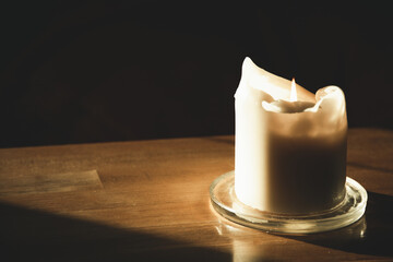Close up shot of a white wax thick candle lit with a flame, burnt to the half, resting on a flat...