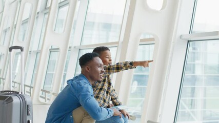Happy young african american dad with preschool son waiting for flight standing at airport and watching planes fly out the window.