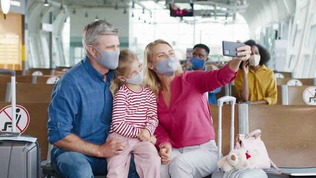 Happy Caucasian family with daughter in protective masks sitting in waiting room of airport before departure and taking selfies on smartphone.