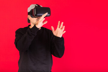 Young man with short beard, wearing black sweatshirt, cap and virtual reality glasses amazed, touching something virtually, on red background. Technology, VR, computing and hobbies concept.