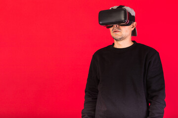 Young man with short beard, wearing black sweatshirt, cap and virtual reality glasses amazed, on red background. Technology, VR, computing and hobbies concept.