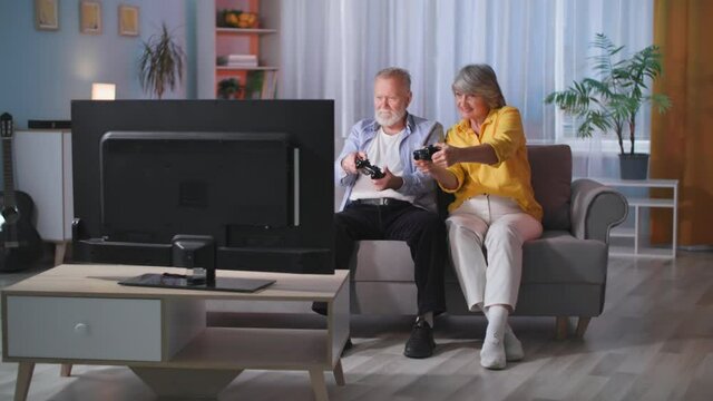 Older Male And Female Gamers Use Modern Technology And Play Video Games On A TV With A Joypad And Game Console While Sitting On A Living Room Couch In The Evening