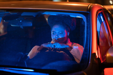 Depressed upset african american young woman sitting behind steering wheel in car at night, having...