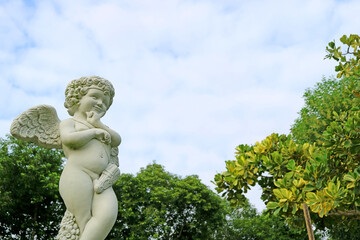 Fototapeta premium Closeup an adorable cupid sculpture in the garden against fluffy cloudy sky