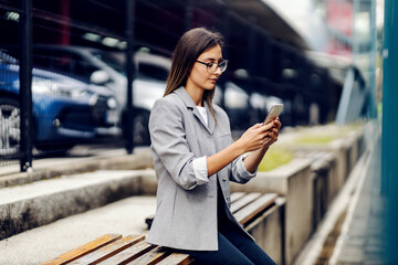 A successful young businesswoman dressed smart casual is sitting outdoors and replying messages to...