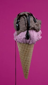 Tasty ice cream cone with two chocolate and strawberry flavors scoops. Pouring syrup topping in waffle cone on pink background. Soft cream, gelato icecream scoop in waffle cone over colorful wallpaper