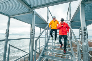 Naklejka na ściany i meble Two cheerfully smiling bright sporty clothes dressed men running down by huge steel industrial stairs with picturesque winter city landscape view. Men's friendship and healthy lifestyle concept photo.