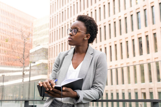 Concentrated Black Female Analyst Writing Notes In Planner On Balcony