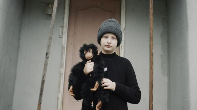 Portrait of a sad girl, standing on the threshold of the house, holding a toy monkey in her hands