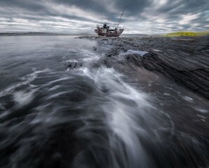 Eine epische Ansicht eines geerdeten Fischereischoners. Das alte Schiff ist mit Rost bedeckt. Düsterer Himmel. Im Vordergrund toben starke Wellen. Barencevo-Meer. Rybachy-Halbinsel. © Olga Rudchenko 