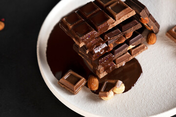 Melted liquid chocolate pours on a plate with pieces of chocolate bars with almond and hazelnut. Close-up.