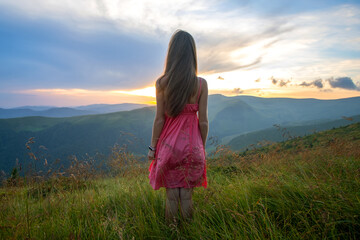 Young happy woman traveler in red dress standing on grassy hillside on a windy evening in summer mountains enjoying view of nature at sunset