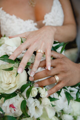 Obraz na płótnie Canvas Newlywed couple holding hands and displaying wedding rings Close up white roses wedding bouquet on the background Two gold wedding rings on the hands of groom and bride White lace dress on background