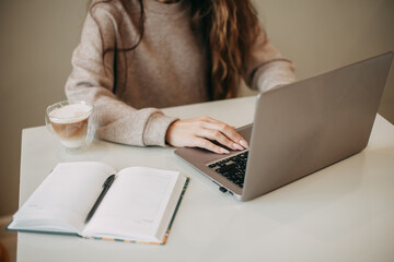 Young brunette woman with long hair uses a laptop at home. There is a mug of coffee and a notebook...