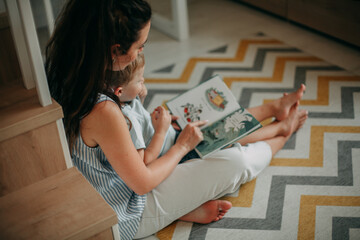 A young girl of 30 years old brunette with long hair in a white shirt with a naked strip and in white jeans at home in the nursery room sits on the floor with her son and reads a book to him