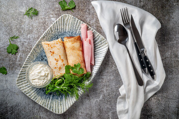 Pancakes filled with ham in a plate with parsley and dill greens and sour cream on a gray stone...