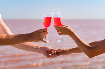 Glasses with a pink drink in the hands of lovers on the background of a pink lake on Valentine's Day. Selective focus on glasses.