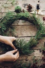 Young woman making Christmas wreath decoration during seasonal workshop, Florist holding Christmas wreath near pinecones, fir tree branches and candle on wood rustic table