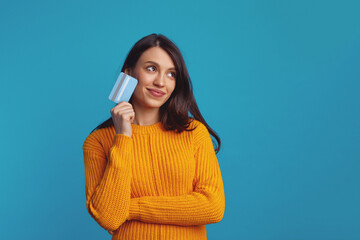 Thoughtful young woman wearing yellow clothes holding plastic credit card near face and looking at...