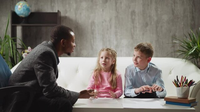 Little blonde girl pupil answers nodding African-American tutor while brother classmate sits on sofa at home English lesson with native speaker slow motion