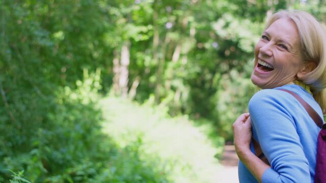 Rear view of mature woman wearing backpack walking ahead of camera along path through trees and looking back over shoulder - shot in slow motion