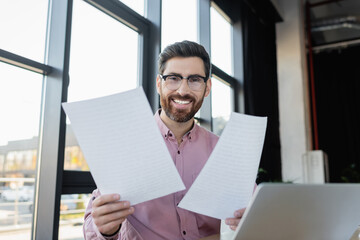 Smiling businessman holding documents near laptop in office.