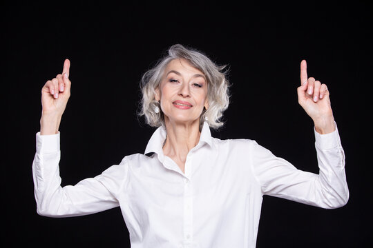 Portrait Of A Modern Happy Senior Woman Pointing Upwards With Two Hands, Isolated On A Black Background