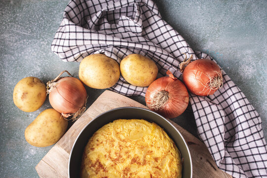 tortilla de patatas en una sart&eacute;n sobre una tabla de cortar de madera, con algunas patatas y cebollas enteras, vista superior
