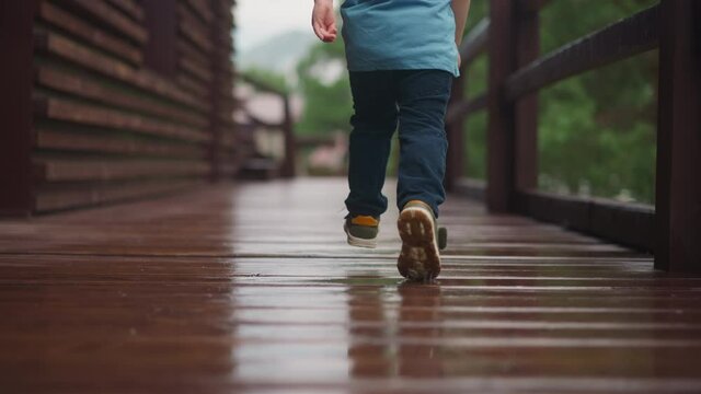 Playful toddler boy in sneakers runs along wet after rain veranda deck with railing at wooden cottage house on spring day close backside view