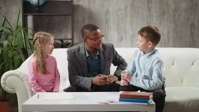 Smart little students answer smiling African-American educator in glasses questions during English lesson with native speaker at home slow motion