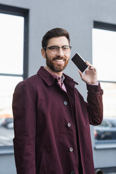 Positive businessman in trench coat looking at camera while holding cellphone with blank screen outdoors.