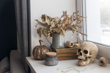 Bouquet of dried autumn leaves in a zinc jug, plastic skull, artificial gilded pumpkin, vintage books decorate the window for Halloween
