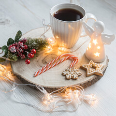 A cup of coffee on a wooden stand, a gingerbread cookie tied with a rope, a garland on the table. Artificial spruce branch with red berries. Breakfast at Christmas. Scandinavian style. Copy space - obrazy, fototapety, plakaty