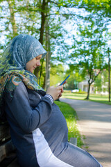 Profile view of the muslim pregnant woman using mobile phone and sitting alone in the park