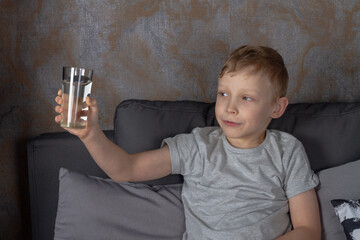 Naklejka premium A caucasian boy drinks water from a glass while sitting on a gray sofa