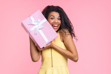Studio image of a beautiful joyful young brunette woman in pastel yellow dress posing isolated over pink wall background holding big present box decorated with bow