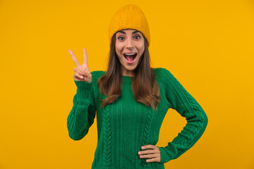 Portrait of young happy woman wearing green knitted sweater and beanie hat posing over bright colored yellow background making victory or peace gesture with excited face expression