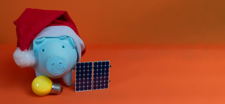 Piggy bank coins with Santa Claus christmas hat, photovoltaic solar panel and lamp isolated on orange background. Concept image.