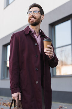 Bearded businessman in trench coat holding bag and paper cup on urban street.