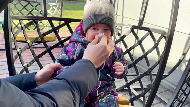 Dad wipes snot to a little Caucasian girl in winter clothes and a gray hat.