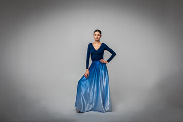 Fototapeta premium Beautiful ballerina stands looking straight into the camera in a long blue dress on a light background