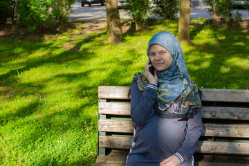 Young muslim pregnant woman talking on the phone while sitting in the park