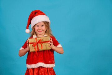Obraz premium cute girl smile Christmas with a gift on the background for the text in a santa costume and a cap. Happy Holidays and present a box with a bow. Copy space. Happy child for the new year. isolate 