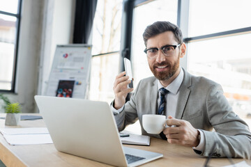 Fototapeta na wymiar Smiling businessman with coffee holding smartphone and looking at camera near laptop.