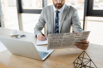 Cropped view of blurred businessman holding newspaper and writing on notebook near laptop in office.