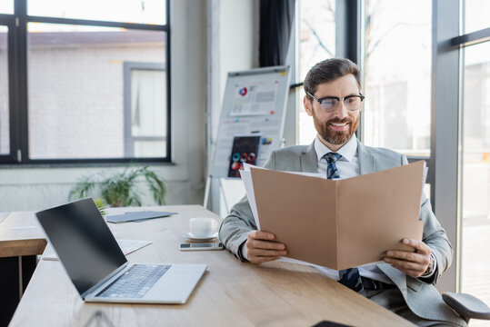 Smiling businessman looking at paper folder near laptop in office.